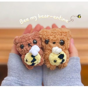 Puede incluir: Dos osos de peluche de crochet marrones que sostienen figuras de abejas de crochet amarillo y negro. Los osos tienen ojos negros y narices blancas. El texto "Bee my bear-entine" está escrito encima de los osos.