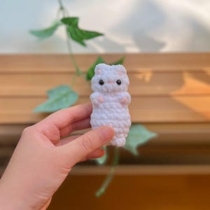 May include: A small, white crocheted animal figure with black eyes and pink accents on its paws and ears. The handmade plush toy is held in a hand, with a wooden background and green leaves visible.