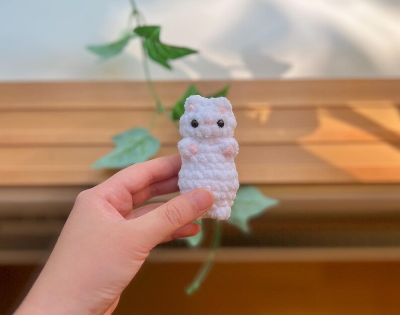 May include: A small, white crocheted animal figure with black eyes and pink accents on its paws and ears. The handmade plush toy is held in a hand, with a wooden background and green leaves visible.