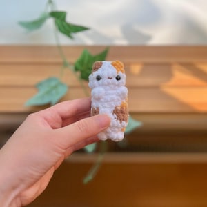 May include: A handmade crochet cat figurine in white, brown, and orange. The small, stuffed cat has a simple design with a rounded body and small facial features. The cat is held in a person's hand against a blurred background of wood and greenery.