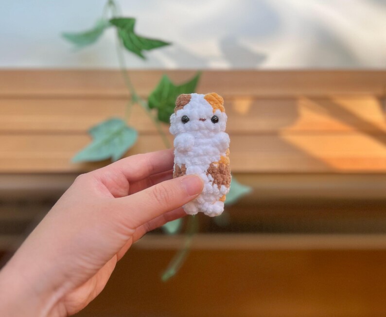 May include: A handmade crochet cat figurine in white, brown, and orange. The small, stuffed cat has a simple design with a rounded body and small facial features. The cat is held in a person's hand against a blurred background of wood and greenery.