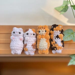 May include: Four handmade crochet cat figures in a row. The cats are white, calico, orange, and brown with black eyes. The figures are standing on a wooden surface with green leaves in the background.