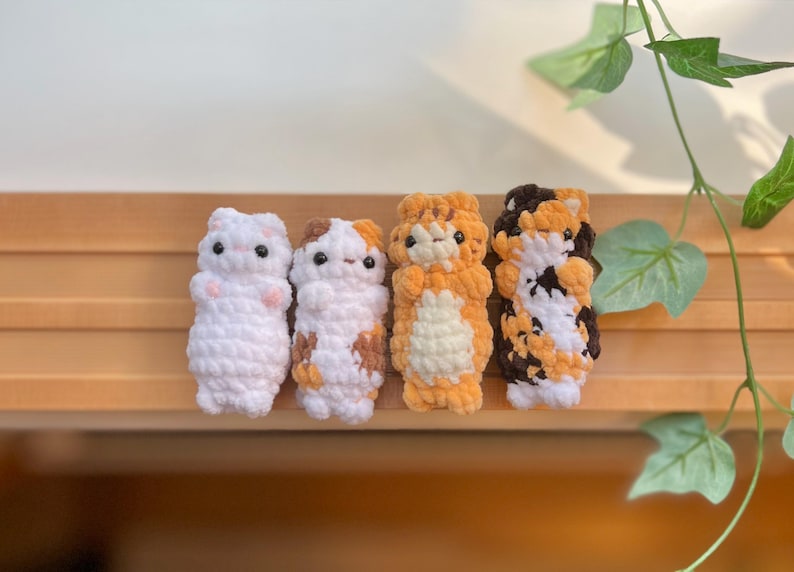 May include: Four handmade crochet cat figures in a row. The cats are white, calico, orange, and brown with black eyes. The figures are standing on a wooden surface with green leaves in the background.