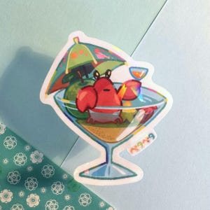 May include: A holographic sticker depicting a cartoon crab in a cocktail glass. The red crab has large claws and wears a gray diaper, under a green umbrella. The glass contains fruit and sand. The sticker has Japanese text.