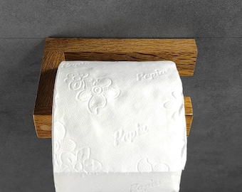 May include: A wooden toilet paper holder with a roll of white toilet paper. The toilet paper has a floral pattern and the word "Papia" printed on it. The holder is made of light brown wood and is mounted on a gray wall.