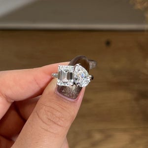 Op de afbeelding: Een zilveren ring met twee grote, heldere edelstenen: een smaragdgeslepen steen en een peervormige steen. De ring wordt getoond tegen een wazige achtergrond, waarbij de schittering van de stenen wordt benadrukt.