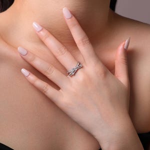 Puede incluir: Un anillo plateado con un dise&ntilde;o entrecruzado, adornado con peque&ntilde;as piedras brillantes. El anillo se luce en una mano con u&ntilde;as cuidadas, sobre un fondo oscuro.