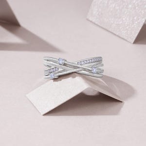 Puede incluir: Un anillo plateado con un dise&ntilde;o entrecruzado. El anillo presenta peque&ntilde;as piedras preciosas transparentes engastadas en la banda. El anillo se muestra sobre una superficie de color claro, con un fondo suave y neutro.