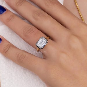 May include: A gold ring featuring a large, rectangular emerald-cut clear gemstone set in a dark, metallic bezel. The stone has prominent facets that reflect light, and the band is a simple, polished gold wire. The ring is worn on a finger.