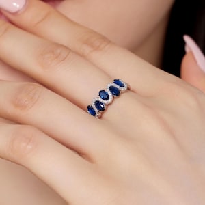 Op de afbeelding: Een zilveren ring met een rij ovale, diepblauwe edelstenen, elk omringd door kleinere, heldere stenen. De ring wordt op een vinger getoond, wat het elegante ontwerp en de schittering benadrukt.