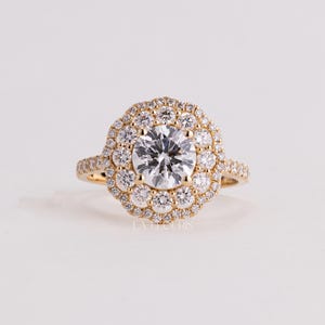 Op de afbeelding: Een gouden ring met een grote ronde briljant geslepen centrale diamant, omringd door een geschulpte halo van kleinere ronde diamanten. De ringband is aan de zijkanten ook bezet met kleine ronde diamanten. De tekst LA ET CURIS is zichtbaar.
