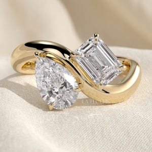 Peut inclure: Une bague en or jaune avec un design bypass, ornée d'un diamant taille poire et d'un diamant taille émeraude côte à côte. L'anneau en or poli entoure les deux pierres facettées transparentes, fixées par des griffes. Le texte LA ELCURIS est visible.