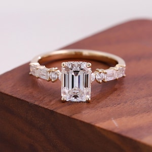 May include: A gold ring with a large, rectangular, clear gemstone as the centerpiece. The band features smaller, rectangular and round gemstones. The ring is set on a wooden surface.