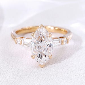 Könnte beinhalten: Ein goldener Verlobungsring mit einem großen, marquiseförmigen Diamanten als Mittelpunkt. Der Ring hat kleinere, rechteckige Diamanten auf beiden Seiten des Hauptsteins, gefasst auf einem zarten Band. Präsentiert auf weißem Hintergrund.