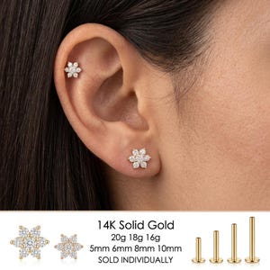 May include: A 14K solid gold flower-shaped stud earring featuring clear stones. The earring is shown in an earlobe and cartilage piercing. Text indicates 20g, 18g, 16g gauges and 5mm, 6mm, 8mm, 10mm lengths for the post, sold individually.