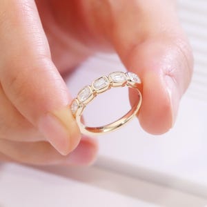 May include: A gold ring featuring a series of rectangular-cut gemstones. The ring's band is a polished gold, and the gemstones are set closely together. The ring is held up by a person's hand, showcasing the ring's design and sparkle.