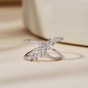 May include: A silver-colored ring with a crisscross design, adorned with numerous small, clear gemstones. The ring features a central cluster of stones and additional stones along the bands, creating a sparkling effect. The ring is set against a neutral background.