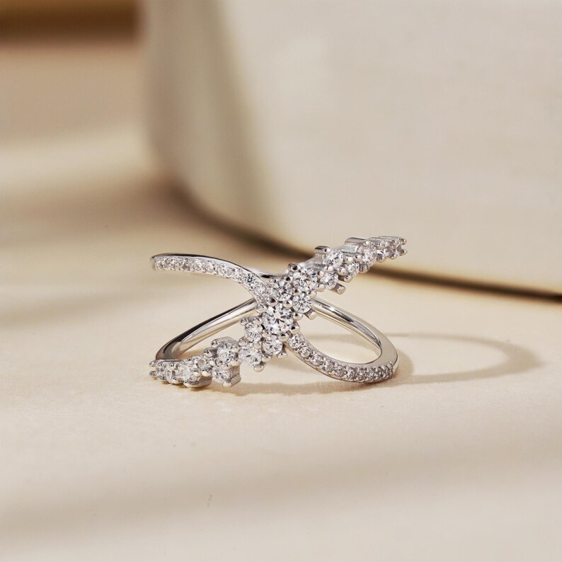 May include: A silver-colored ring with a crisscross design, adorned with numerous small, clear gemstones. The ring features a central cluster of stones and additional stones along the bands, creating a sparkling effect. The ring is set against a neutral background.