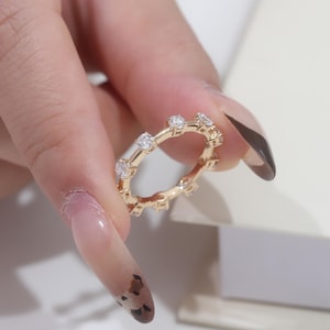 May include: A gold-colored ring with a series of evenly spaced, clear gemstones. The ring is held between fingers, showcasing its delicate design and sparkling stones. The ring's band has a repeating pattern of small, decorative elements.