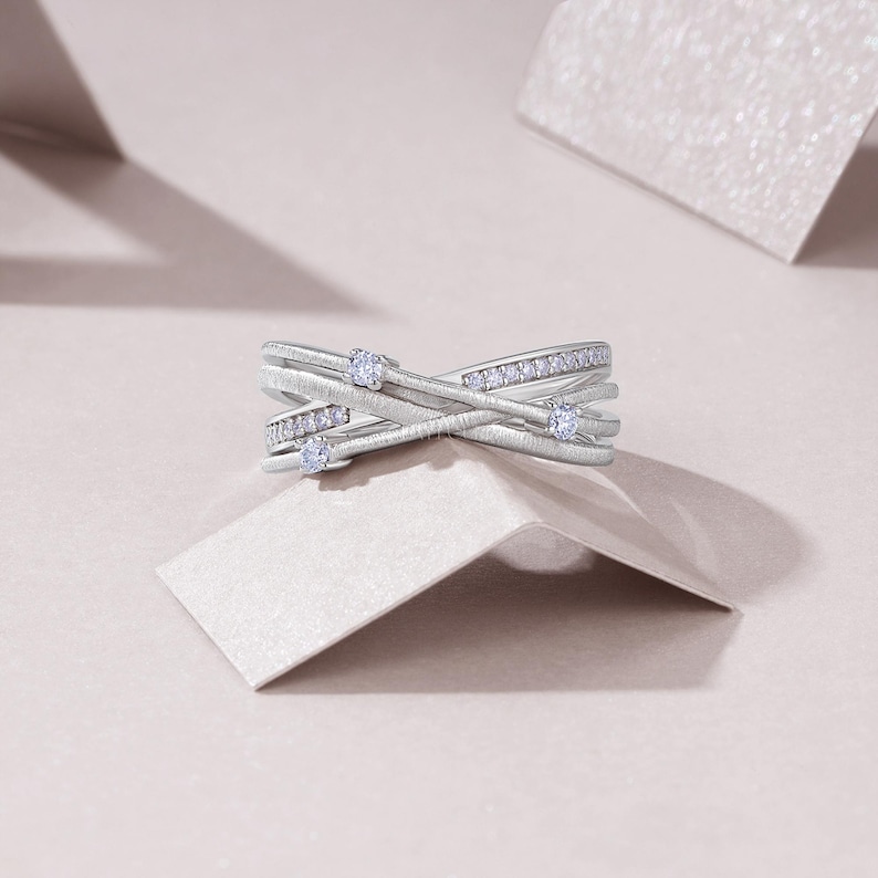 Puede incluir: Un anillo plateado con un dise&ntilde;o entrecruzado. El anillo presenta peque&ntilde;as piedras preciosas transparentes engastadas en la banda. El anillo se muestra sobre una superficie de color claro, con un fondo suave y neutro.