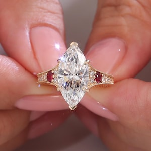 Könnte beinhalten: Verlobungsring aus Gold mit einem Diamanten im Marquiseschliff. Der Ring hat einen großen, klaren Diamanten als Mittelpunkt, flankiert von kleineren roten Edelsteinen und Akzentdiamanten. Das Band hat detaillierte Muster.