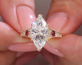 以下が含まれることがあります： ゴールドの婚約指輪。マーキス型のダイヤモンドが特徴で、中央に大きなクリアダイヤモンド、両側に小さな赤い宝石とアクセントダイヤモンドが配置されています。バンドには複雑なディテールが施されています。