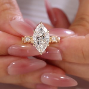 May include: A marquise-cut diamond engagement ring with a gold band. The ring features a large, clear diamond as the centerpiece, set with prongs. The band has a delicate, ornate design with smaller accent diamonds.