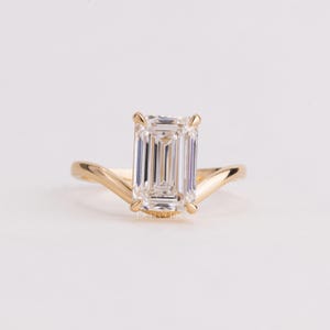 May include: A gold ring featuring a large, rectangular emerald-cut clear gemstone set in four prongs. The band has a distinct V-shaped curve that meets the base of the stone setting, creating a sleek, polished appearance with a metallic finish.