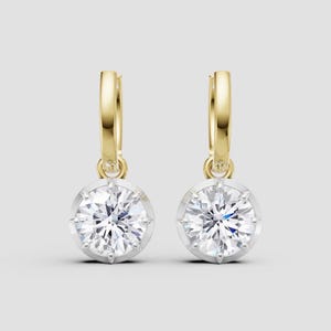 Puede incluir: Un par de pendientes de aro dorados con colgantes redondos de piedras preciosas transparentes. Las piedras están engastadas en un bisel plateado con un diseño de estrella. Fondo gris claro.