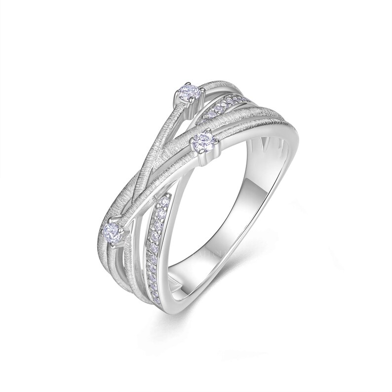 Puede incluir: Un anillo plateado con un dise&ntilde;o fluido e entrelazado. El anillo presenta tres piedras claras m&aacute;s grandes y una fila de piedras m&aacute;s peque&ntilde;as. La banda tiene una apariencia texturizada y cepillada, creando un aspecto moderno y elegante. El anillo est&aacute; sobre un fondo blanco.