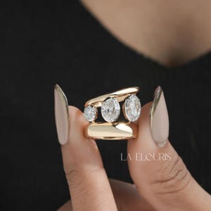 May include: A gold ring featuring three oval-cut clear stones set in a horizontal row between two smooth gold bands. The ring has a modern open-space design. The text LA ELOURIS is visible in the lower right corner of the image.