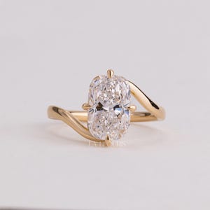 May include: A gold ring featuring a single elongated cushion-cut diamond set in a four-prong setting. The band has a bypass design that curves around the central stone. The text LA ELOURIS is visible at the base of the ring setting.