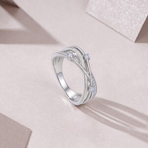 Puede incluir: Un anillo plateado con un dise&ntilde;o tejido. El anillo presenta tres piedras claras m&aacute;s grandes y piedras de acento m&aacute;s peque&ntilde;as. La banda tiene un acabado texturizado y el anillo est&aacute; sobre un fondo rosa p&aacute;lido.