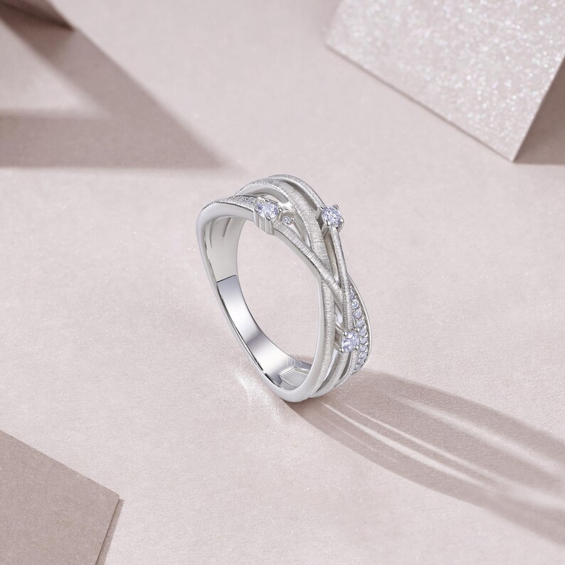 Puede incluir: Un anillo plateado con un dise&ntilde;o tejido. El anillo presenta tres piedras claras m&aacute;s grandes y piedras de acento m&aacute;s peque&ntilde;as. La banda tiene un acabado texturizado y el anillo est&aacute; sobre un fondo rosa p&aacute;lido.