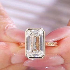 以下が含まれることがあります： 大きな長方形の透明な宝石が付いたゴールドリング。 宝石は金のベゼルにセットされています。 リングは、ピンクのマニキュアを塗った2つの手で持たれています。