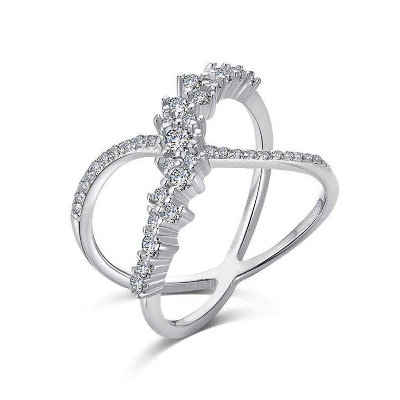 May include: A silver-colored ring with a unique crisscross design. The ring features a row of clear, sparkling stones set in a vertical line, with smaller stones outlining the curved bands. The ring is set against a white background.
