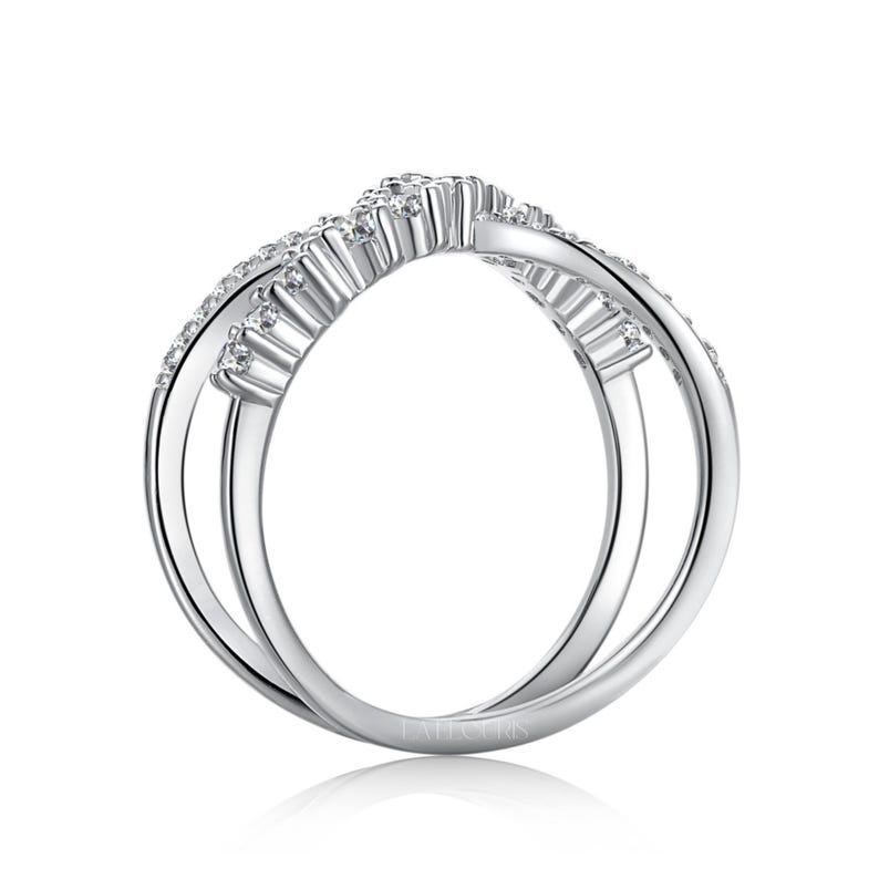 May include: A silver-colored ring with a unique, intertwined design. The ring features a curved top section adorned with small, sparkling stones, likely diamonds or cubic zirconia. The band has a double-loop design, creating an elegant and modern look. The ring is a piece of jewelry.