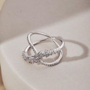May include: A silver-toned ring with an intertwined design. The ring features a row of small, clear gemstones along one band and a cluster of larger gemstones at the center. The ring is set against a neutral background.