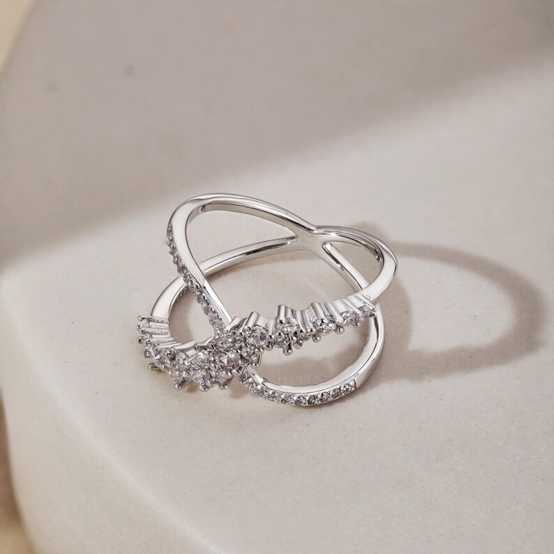 May include: A silver-toned ring with an intertwined design. The ring features a row of small, clear gemstones along one band and a cluster of larger gemstones at the center. The ring is set against a neutral background.