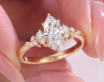以下が含まれることがあります： 中央にマーキス型のダイヤモンドをあしらったゴールドの婚約指輪。小さなラウンドダイヤモンドが両側に配置されています。リングは繊細なバンドとメインストーンの両側にクラスターデザインがあります。リングは人の指で持たれています。