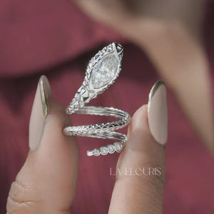May include: A silver-toned snake-shaped ring featuring a marquise-cut clear stone at the head and a textured, coiled band that wraps around the finger. The tail end is accented with a row of small, round clear stones. The text LA ELOURIS is visible.