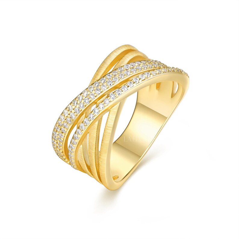 May include: A gold-colored ring with a crisscross design. One band is encrusted with small, clear gemstones. The ring has a polished finish and is set against a white background. The ring's design features multiple interwoven bands.