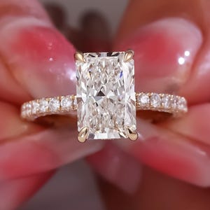 May include: A radiant-cut diamond engagement ring. The ring features a large, rectangular, clear diamond set in a gold band. The band is adorned with smaller diamonds, adding sparkle. The ring is held against a soft pink background.