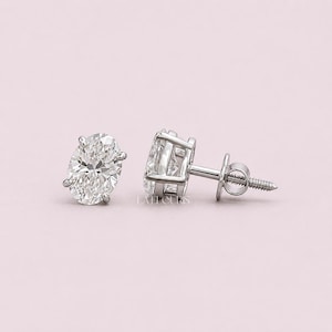 May include: A pair of oval-cut diamond stud earrings. The diamonds are set in a four-prong setting with a screw-back post. The earrings are displayed on a light pink background.