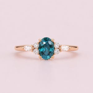 May include: A rose gold ring featuring a teal oval-cut gemstone as the centerpiece, flanked by smaller white gemstones. The ring's band is slender, and the setting includes small gold accents. The overall design is elegant and delicate.