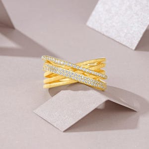 May include: A gold-colored ring with a crisscross design. One band is encrusted with small, clear gemstones. The ring is set against a neutral background with geometric shapes, highlighting the jewelry's design and sparkle.