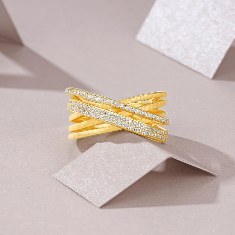 May include: A gold-colored ring with a crisscross design. One band is encrusted with small, clear gemstones. The ring is set against a neutral background with geometric shapes, highlighting the jewelry's design and sparkle.