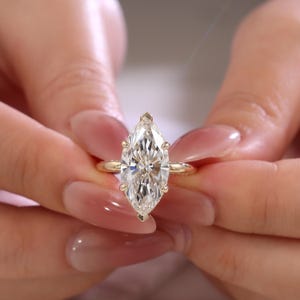 May include: A marquise-cut clear gemstone ring set in a polished yellow gold band. The center stone is held by six prongs, with a delicate milgrain texture along the metal setting. The band is smooth and circular, showcasing a classic design.