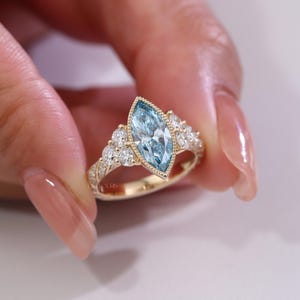 May include: A gold ring featuring a marquise-cut, light blue gemstone as the centerpiece, flanked by smaller, round, clear gemstones. The ring's band is also adorned with small, clear gemstones, creating a sparkling effect. The ring is held up by a person's hand.