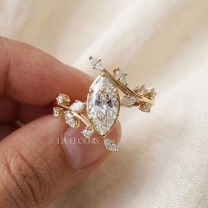 May include: A gold ring featuring a large marquise-cut center stone surrounded by smaller marquise and round-cut stones arranged in a branch-like pattern. The text LA ELOURIS is visible on the band. The design incorporates multiple clear stones.